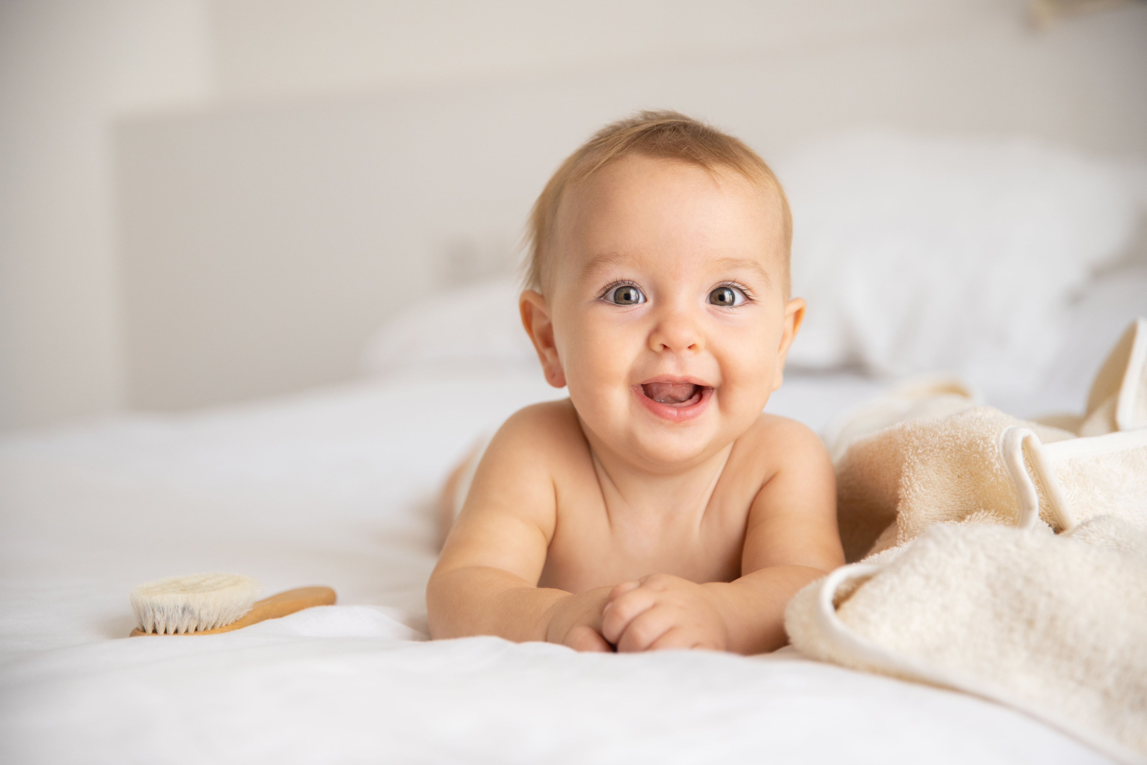 Smiling baby on white bedding with a wooden baby brush and towel — natural baby care